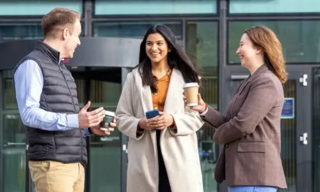 Three students outside the AMBS building chatting and drinking coffee