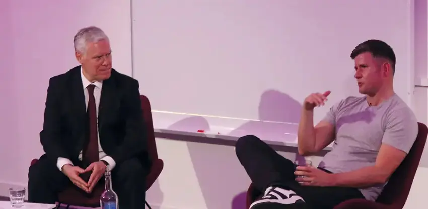 Tom Beahon and Ian King sat in front of some small tables having a conversation at the front of a conference room