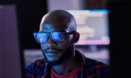 A man working on a computer in dark lighting, with the screen reflecting in his glasses.