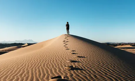 A woman walking up a hill in a desert, leaving foot steps behind her.