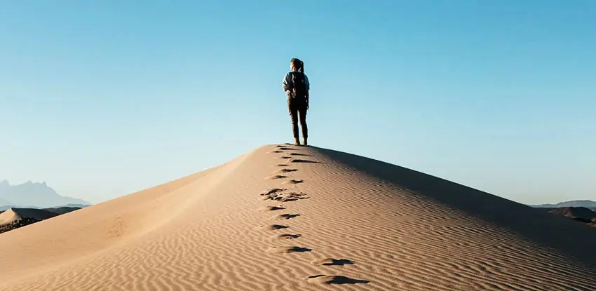 A woman walking up a hill in a desert, leaving foot steps behind her.