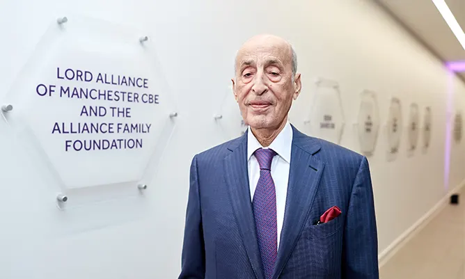 Photograph of Lord David Alliance standing beside his donor plaque at Alliance Manchester Business School